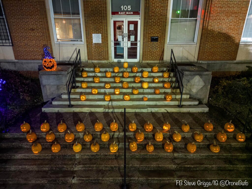 Rows of carved pumpkins with varied designs are displayed on the steps leading to a brick building entrance.