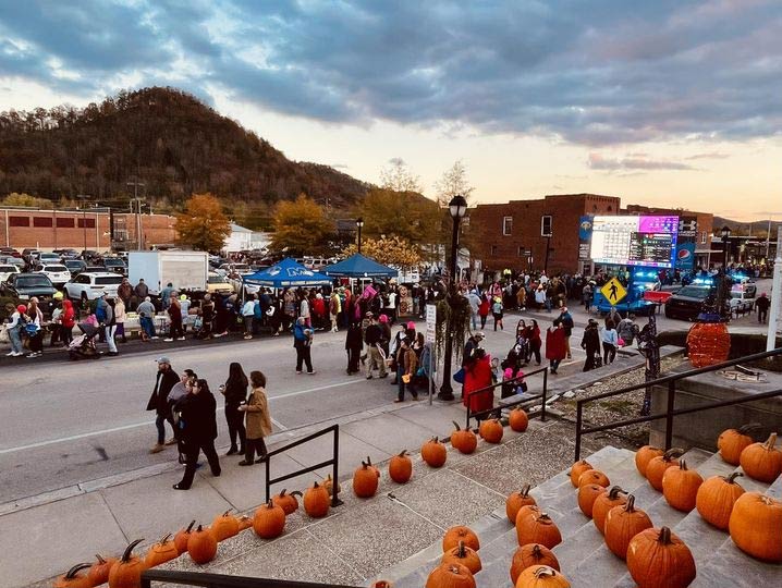 Street scene with a crowd gathered, lined pumpkins, and a backdrop of hills and buildings at dusk.