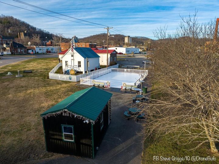 Aerial view of a small fenced ice rink beside a building with a green roof in a grassy area with leafless trees and surrounding structures.