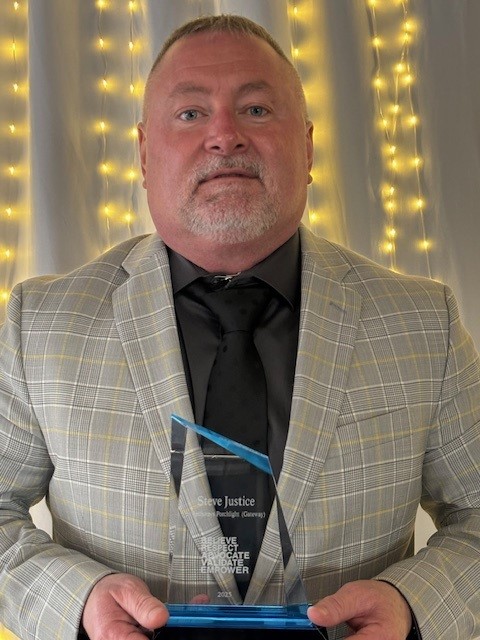 A person in a plaid suit holds a glass award with a curtain and string lights in the background.