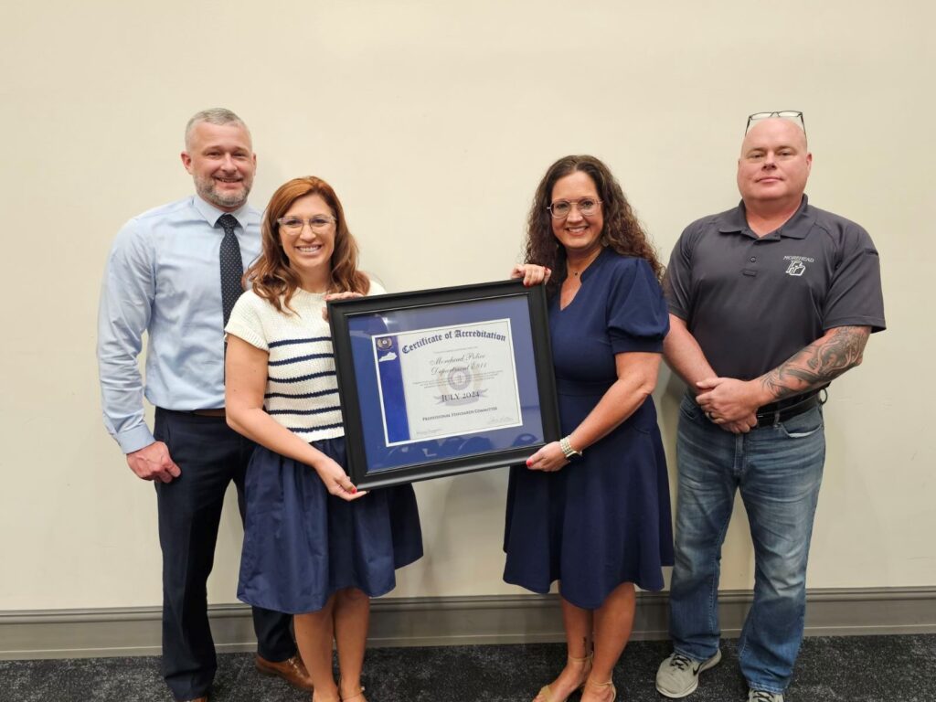 Four people stand together, two holding a framed certificate of accreditation, against a plain background.