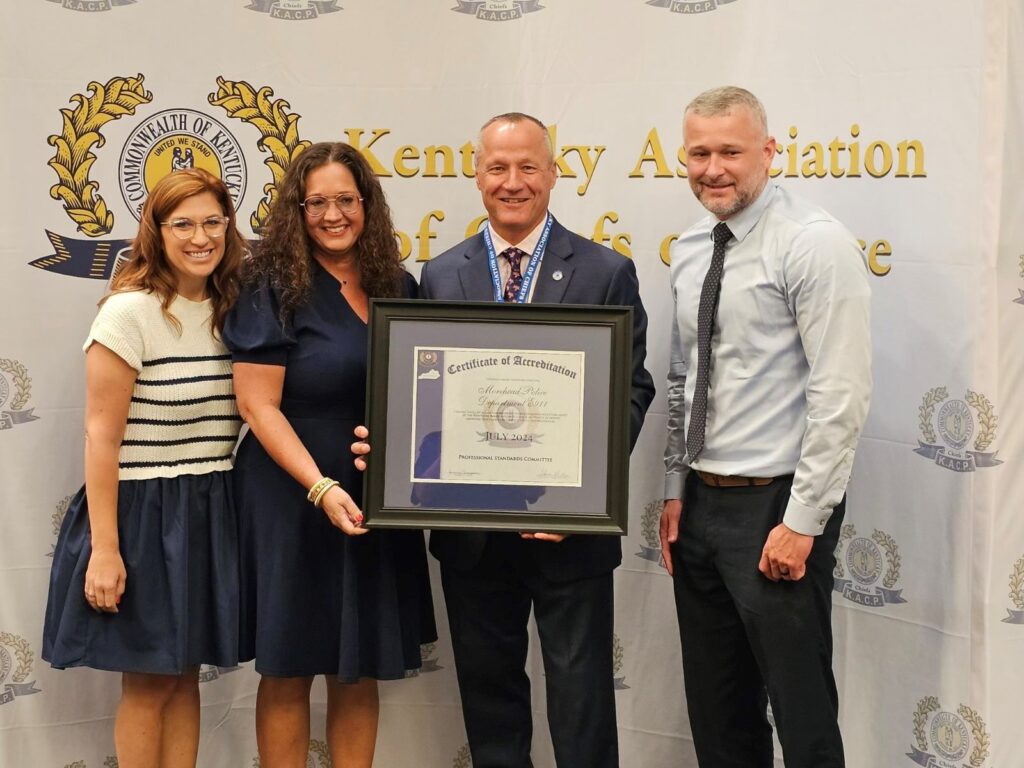 Four people stand together, smiling, holding a framed certificate in front of a Kentucky Association of Chiefs of Police banner.