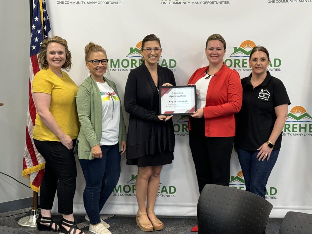 Five people stand together indoors, holding a certificate, with an American flag and Morehead banner in the background.