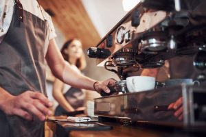 Barista operating an espresso machine with a white cup placed under the portafilter as another person stands in the background.