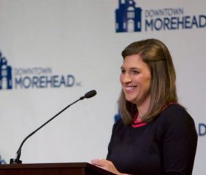 Person standing at a podium speaking into a microphone with a Downtown Morehead backdrop.