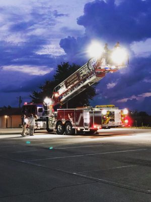A fire truck with an extended ladder and lights illuminates a parking lot at dusk, with two firefighters on the ladder.