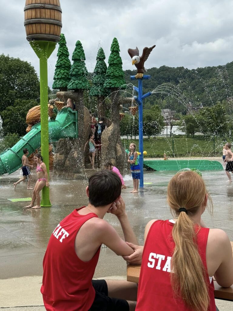 Children play in a water park with slides and water features, supervised by staff in red shirts.