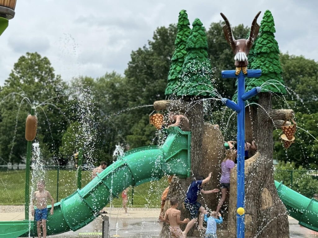 Children play in a splash park featuring water slides and tree-themed structures with water sprays.