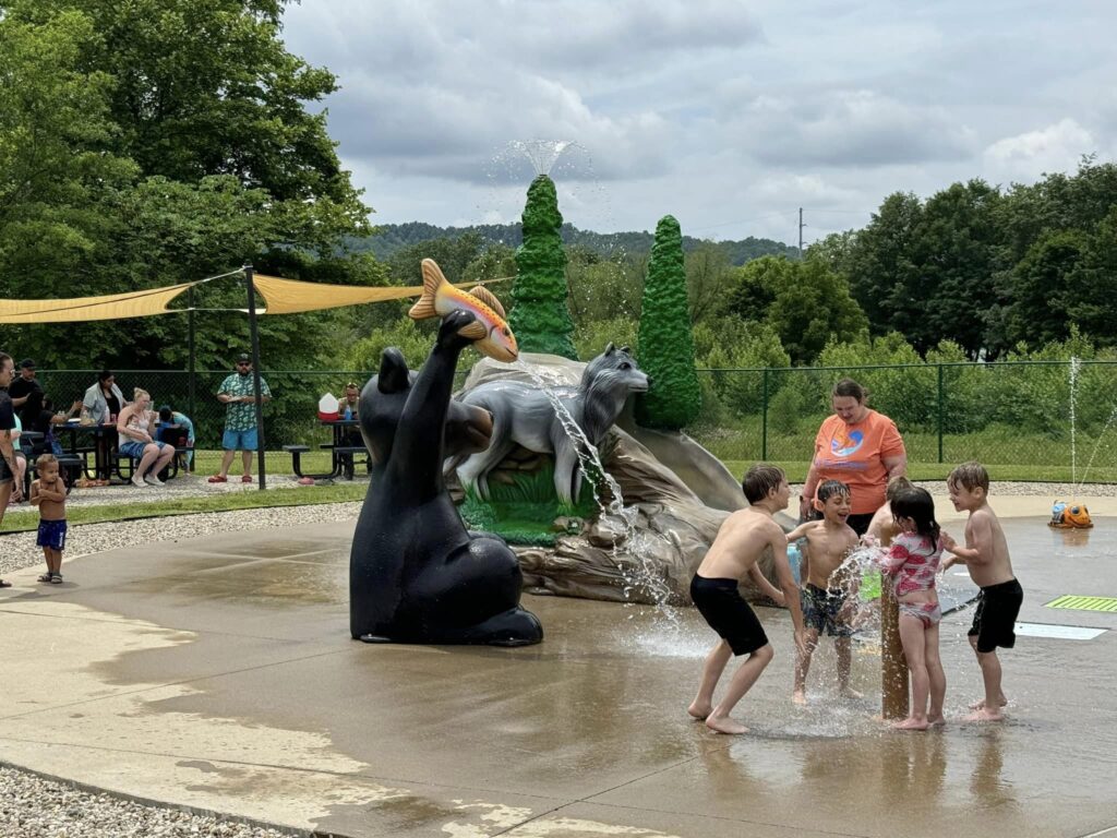 Children play in a splash pad area with animal sculptures and water features, surrounded by trees and picnic tables.