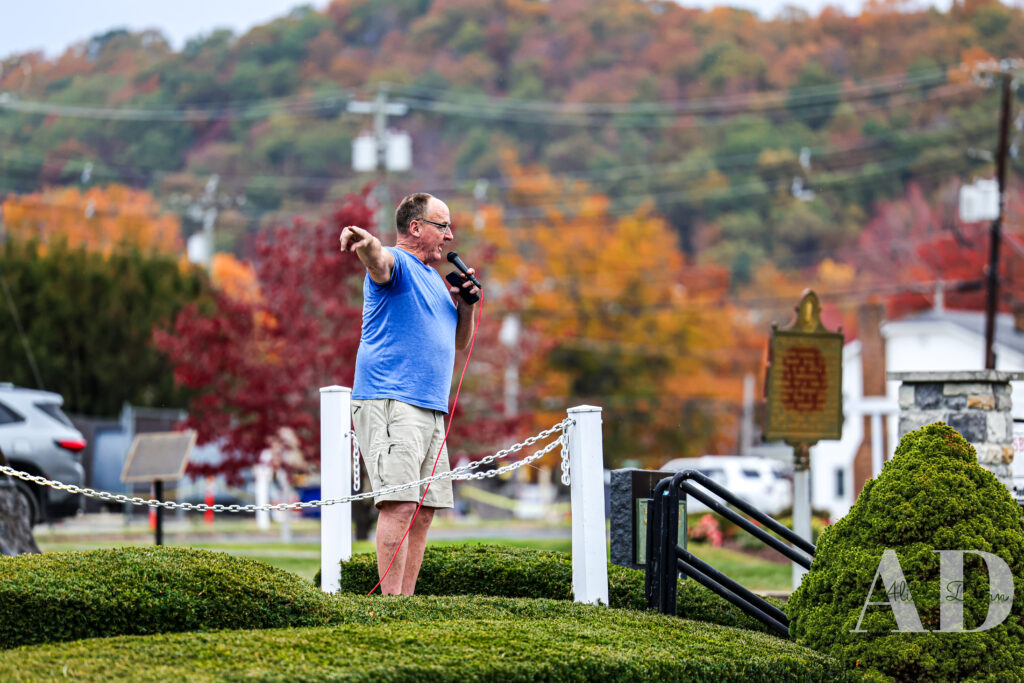 A person stands on a landscaped lawn, holding a microphone and pointing, with autumn foliage and buildings in the background.