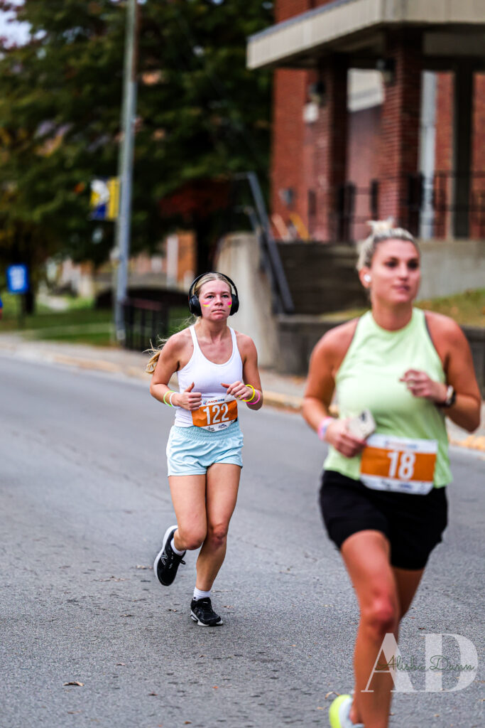 Two runners with numbered bibs participating in a road race, one in focus wearing headphones.