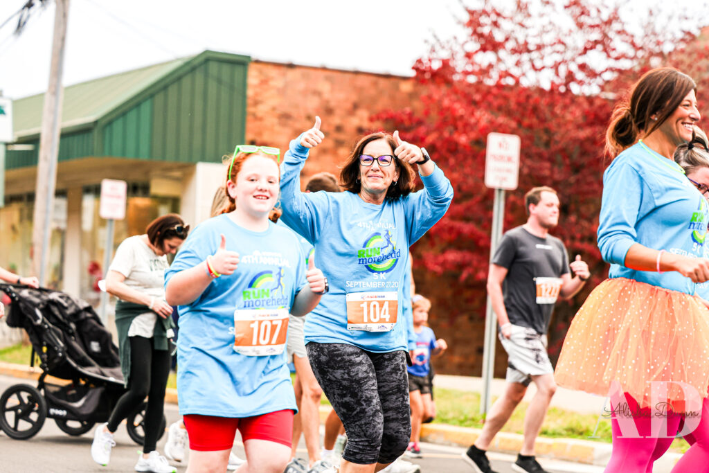 Participants in a community run wearing numbered bibs, walking along a street with autumn trees in the background.