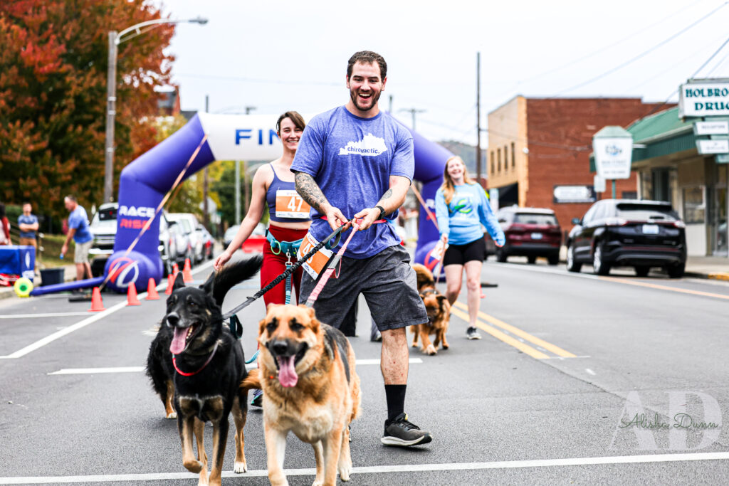 People walk with dogs on leashes down a street near a finish line sign during an outdoor event.