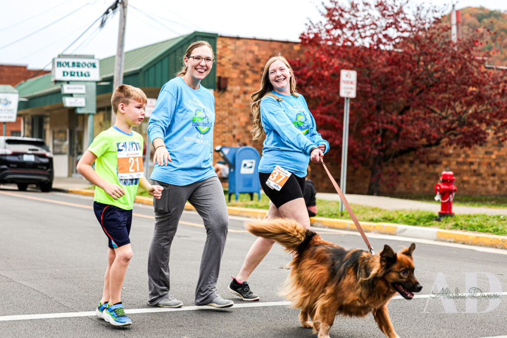Three people and a dog walk in a street event wearing numbered race bibs, with buildings and trees in the background.