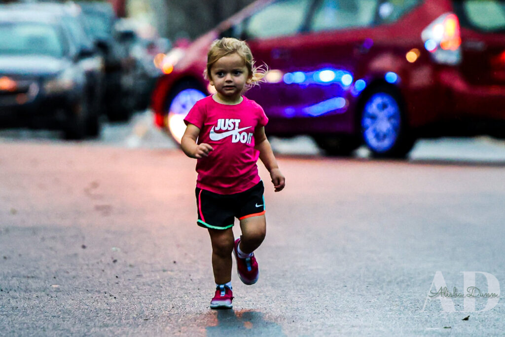 Child in a pink shirt and black shorts running on a street with parked cars in the background.