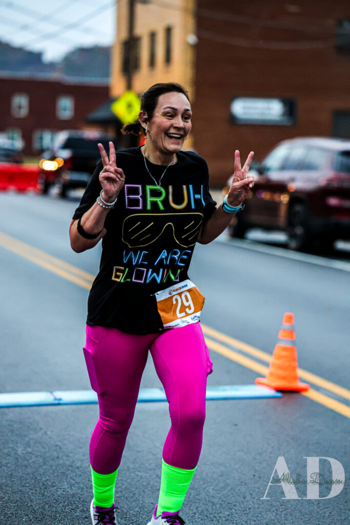 A runner wearing a race bib, pink leggings, and a black shirt makes peace signs while participating in a street event.