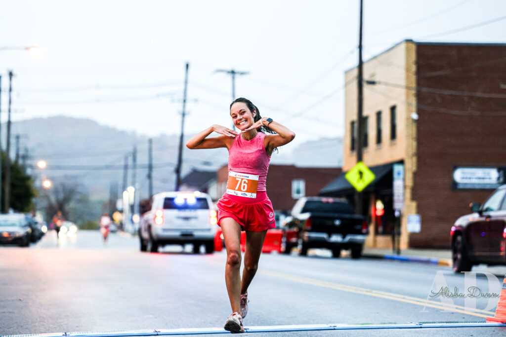 A runner wearing a pink outfit and bib number 76 smiles while crossing a street during a race, with vehicles in the background.
