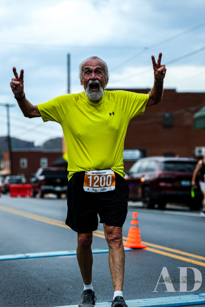 A runner in a yellow shirt and race bib 1200 gestures at the finish line on a city street.
