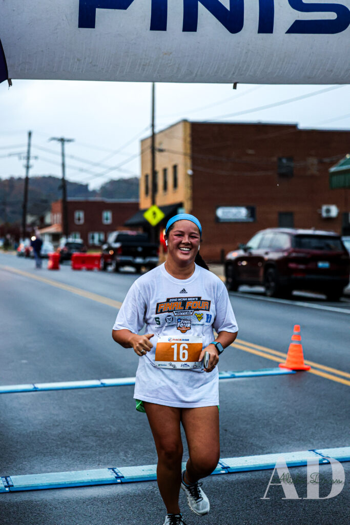 Runner crosses the finish line on a street during a race, wearing a numbered race bib and a headband.