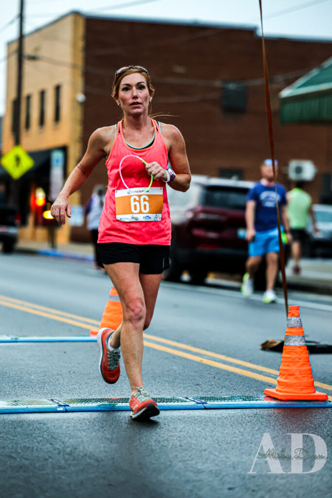 A runner wearing a pink tank top and black shorts crosses a street race finish line marked with traffic cones.