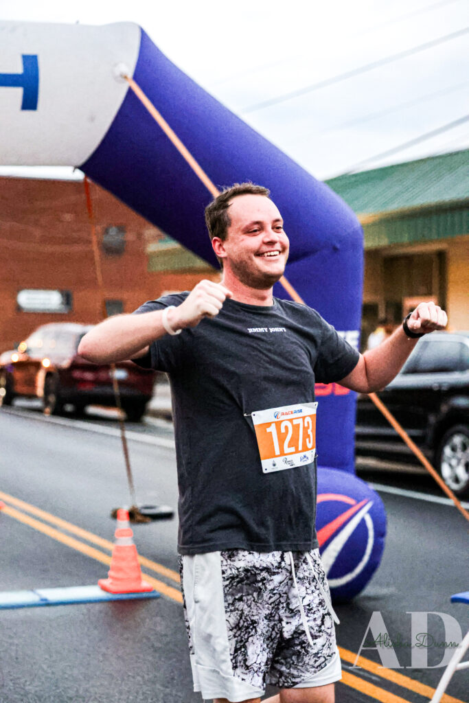 Runner crossing a finish line with an orange bib number 1273, smiling under a blue race arch on a street.
