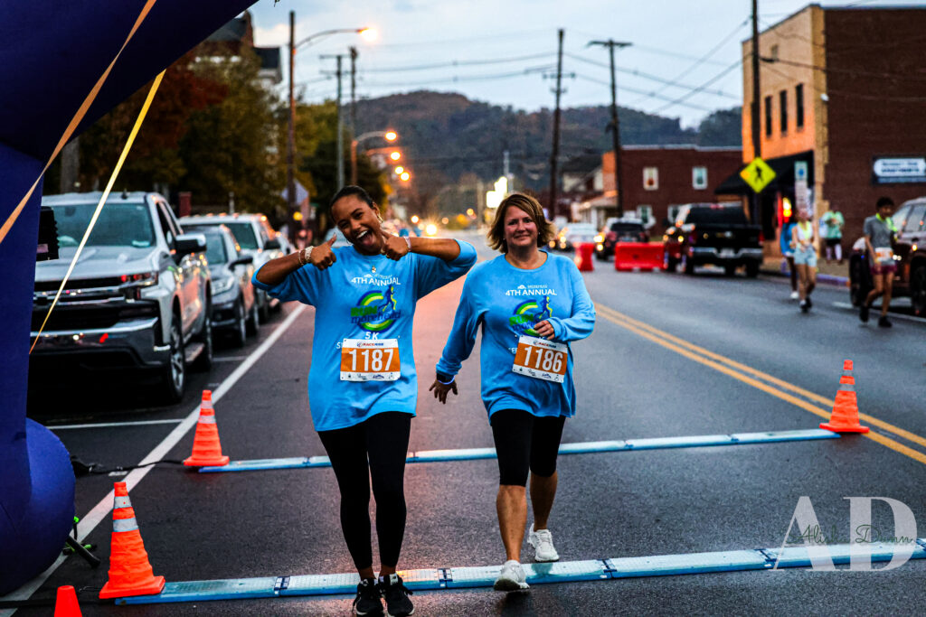 Two runners crossing a finish line during a street race, wearing blue event shirts and numbered bibs.