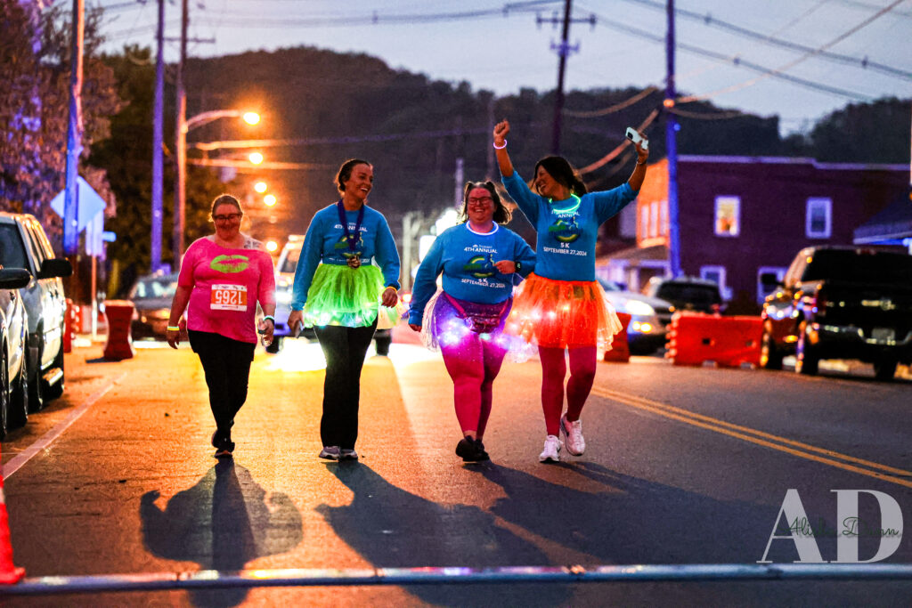 Four people in colorful clothing walk down a dimly lit street at night, with overhead streetlights illuminating the scene.