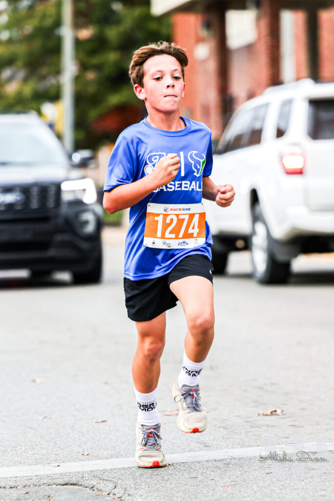 A young runner in a blue shirt and black shorts jogs on a city street, wearing race bib number 1274.