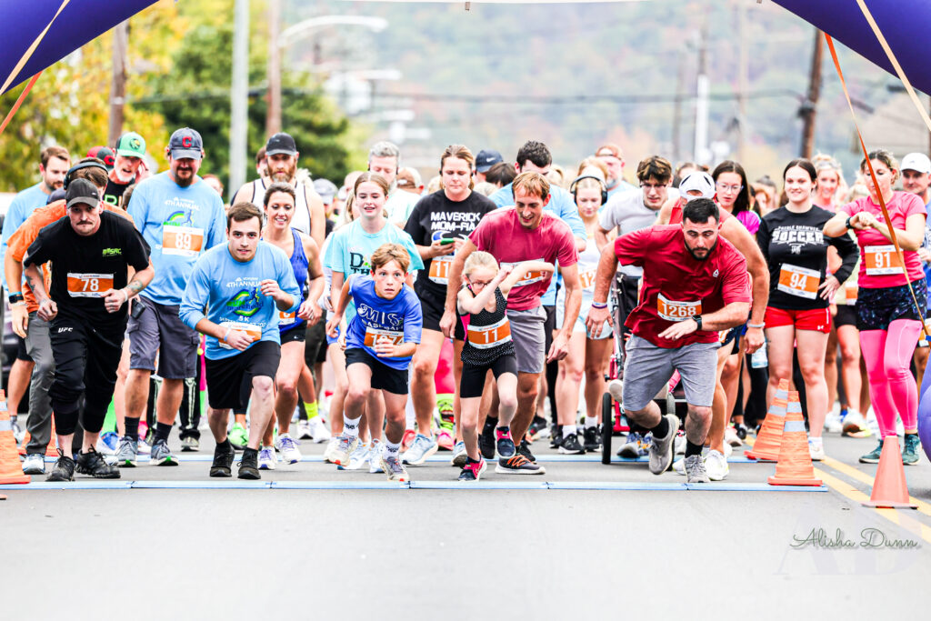 Participants begin a road race, crossing the starting line under inflatable archway, with orange cones on either side.