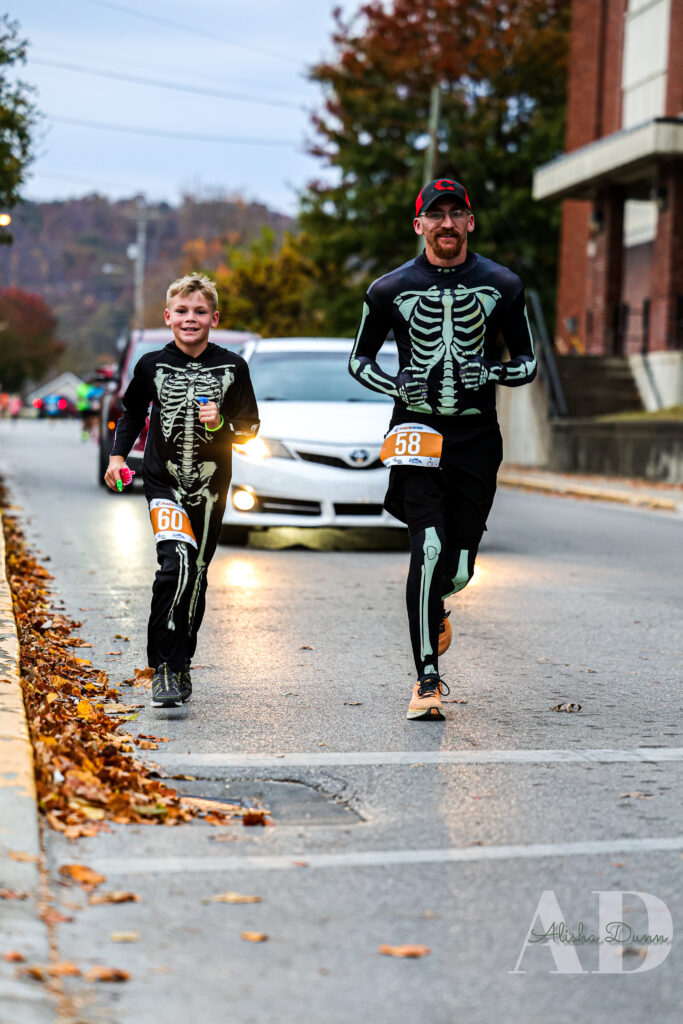Two people in skeleton costumes run in a street race with fallen leaves along the sidewalk.