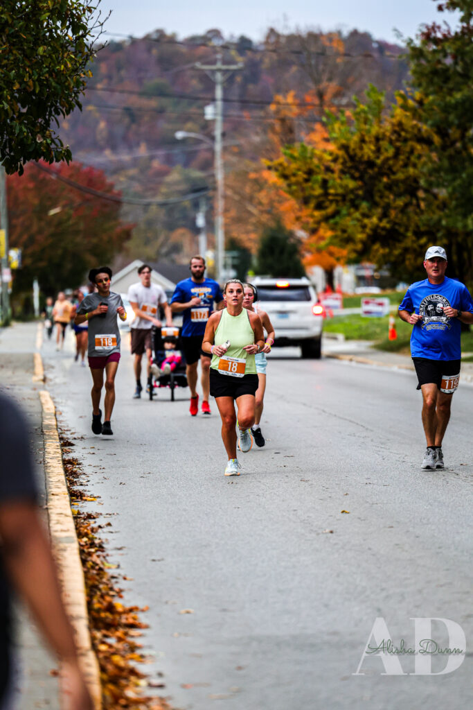 Runners participate in a street race with autumn trees in the background, some wearing numbered bibs.