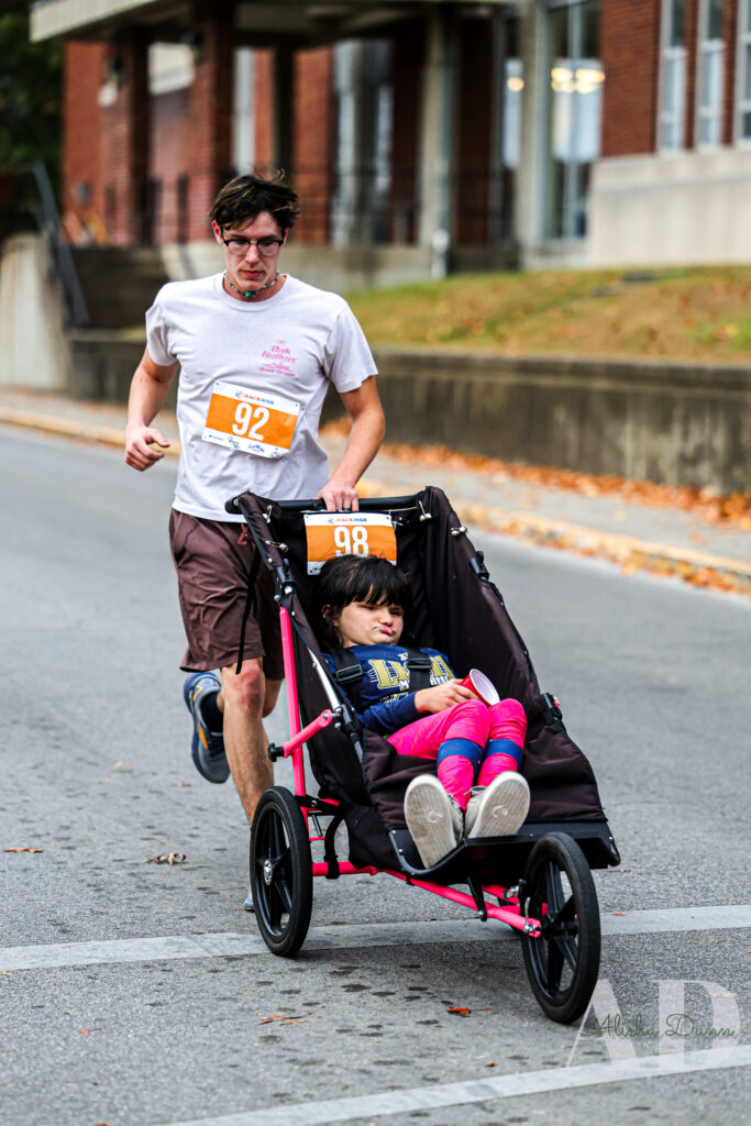 A man runs on a street pushing a stroller with a child. Both wear race numbers; buildings are in the background.