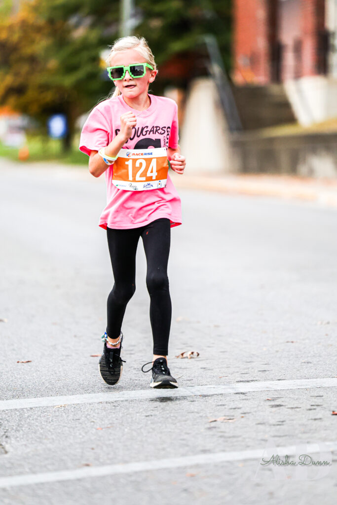 A young runner wearing a pink shirt and green sunglasses participates in a road race wearing bib number 124.