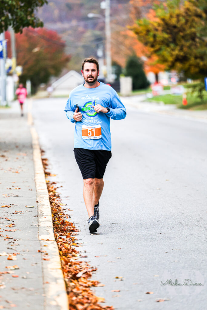 Runner in a blue shirt and black shorts participating in a road race, with autumn leaves along the roadside.