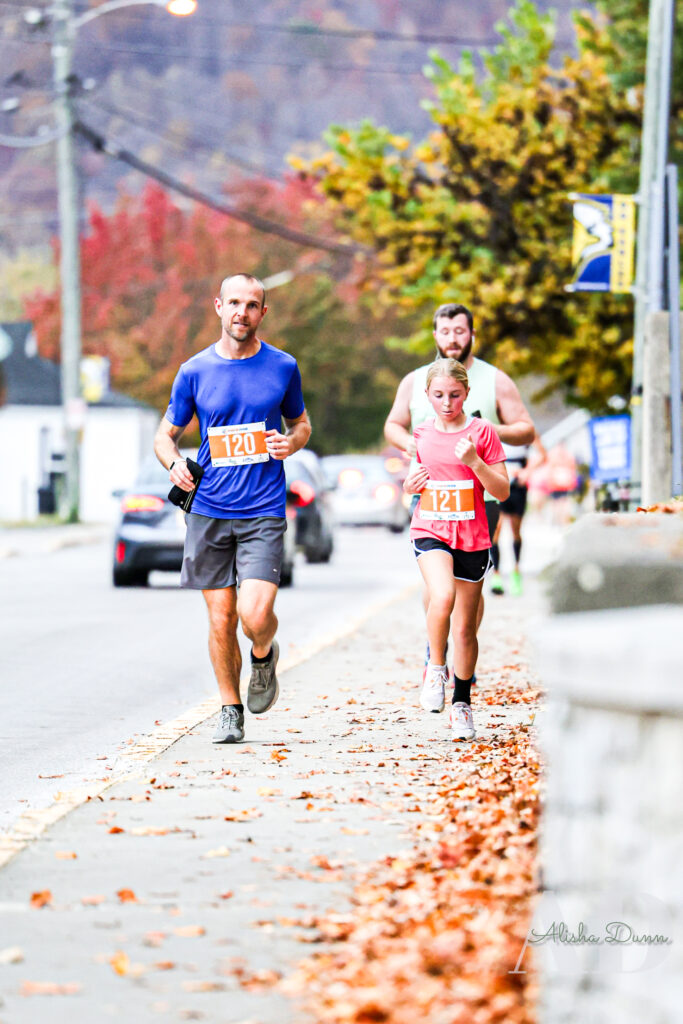 Runners with numbered bibs jog along a sidewalk lined with autumn leaves, with cars and trees in the background.