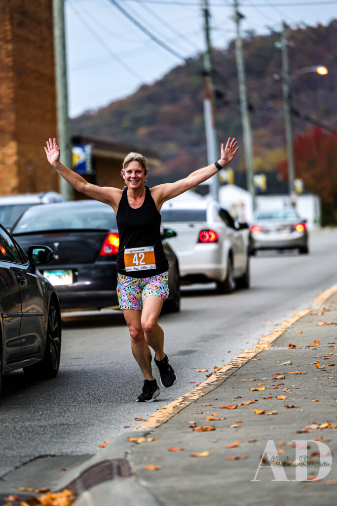 Runner with arms raised jogging along a street, wearing a numbered bib, with cars and autumnal trees in the background.