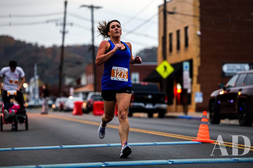 Runner in a blue tank top crosses a race finish line on a city street, with traffic cones and buildings in the background.