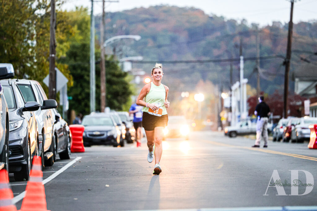 Runner in a race on a street lined with parked cars and traffic cones, with trees and hills in the background.