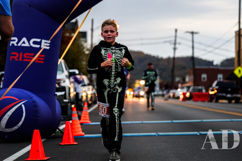 Child in a skeleton costume crosses the finish line of a race, with an inflatable archway and traffic cones in the background.