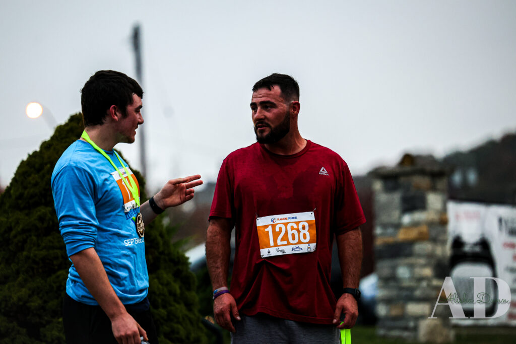 Two men with race bibs discuss outdoors; one wears a medal, the other in a red shirt with a bib labeled 1268.