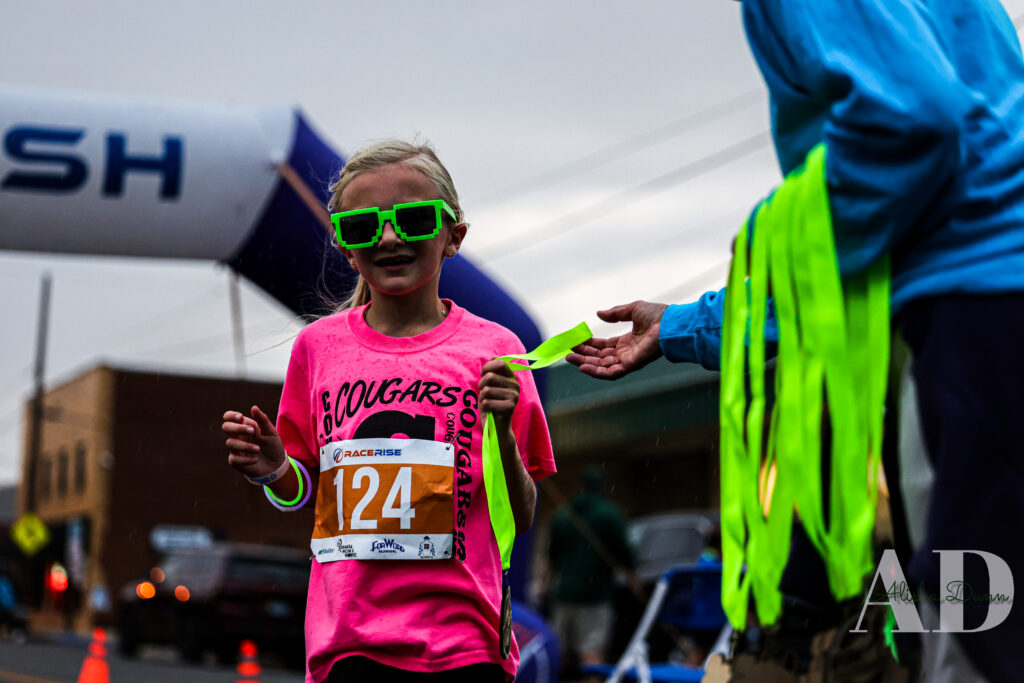 A young participant wearing a number 124 bib receives a finisher ribbon at a race event finish line.