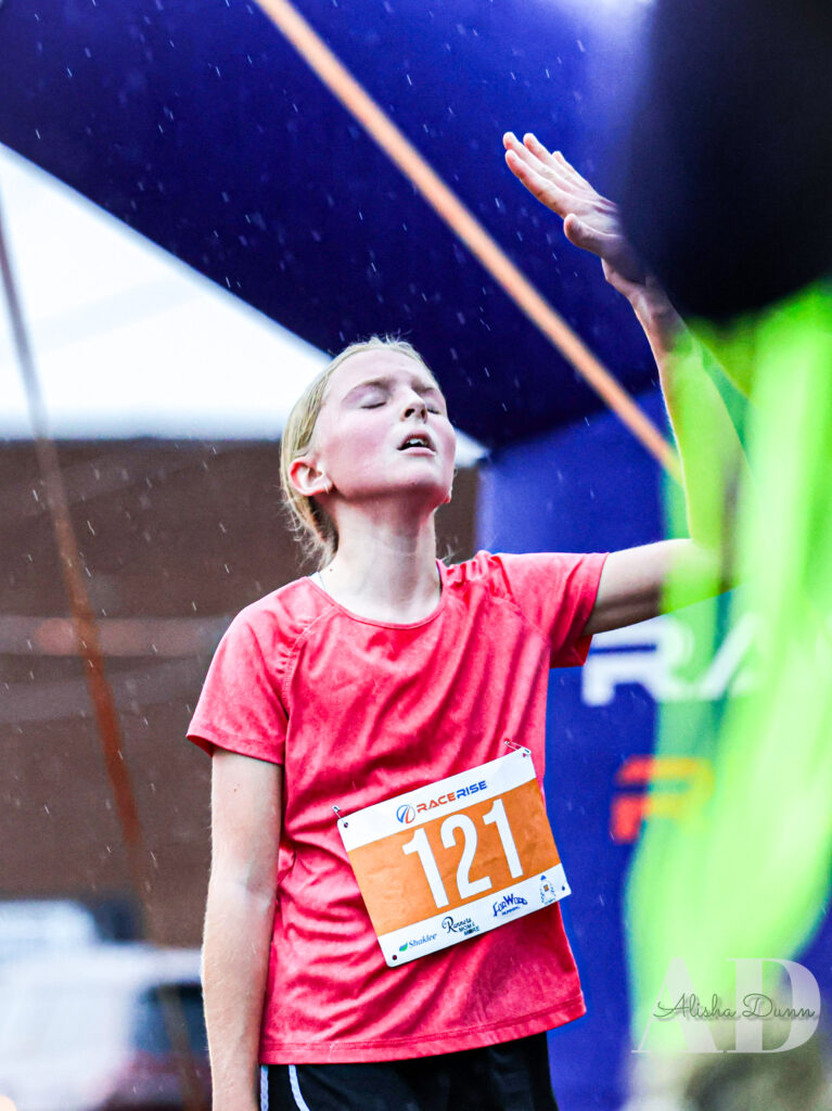 A runner wearing a red shirt and race bib number 121 stands with eyes closed, reaching upward during a rainy event.