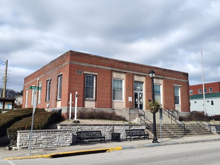 Photograph of the historic red-brick former Morehead Police Station at 405 E. Main Street, featuring stone foundation walls, front steps, a lamp post, and a street sign under a cloudy sky.