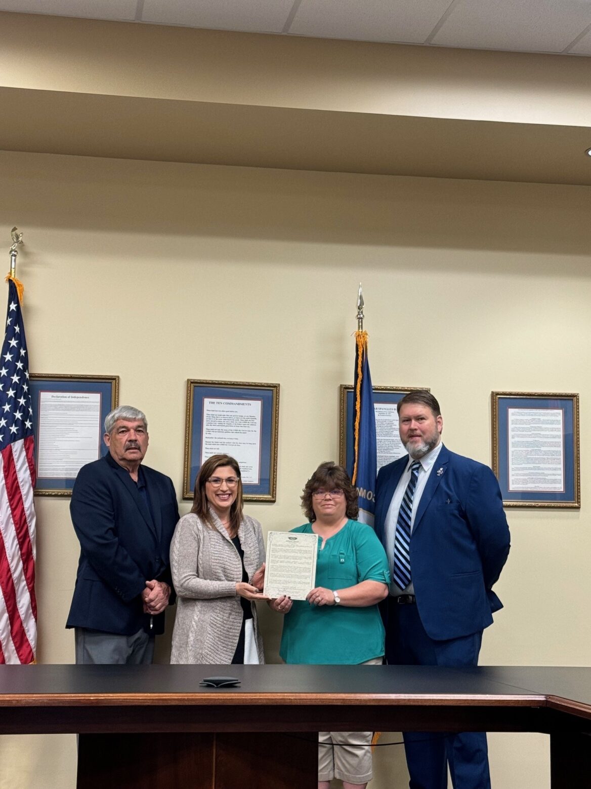 Four city officials stand behind a dark wood conference table against a beige wall lined with framed proclamations. From left to right: an older man in a dark blazer, a smiling woman in a light gray cardigan, a woman in a teal blouse holding a printed proclamation alongside the woman in the cardigan, and a bearded man in a navy suit and striped tie. To the far left is an American flag and to their right a Kentucky state flag.