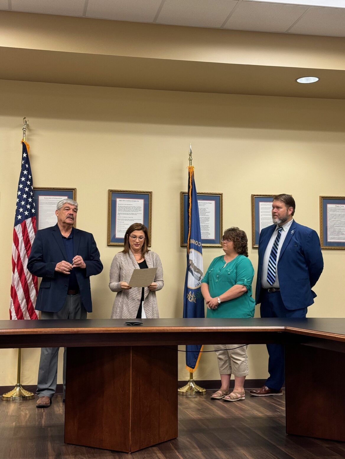 The same four officials behind the conference table. The woman in the gray cardigan, now second from left, holds and reads aloud from the proclamation while the man in the dark blazer (far left), the woman in teal (second from right), and the man in the navy suit (far right) listen. The flags and framed documents remain visible on the beige wall behind them.