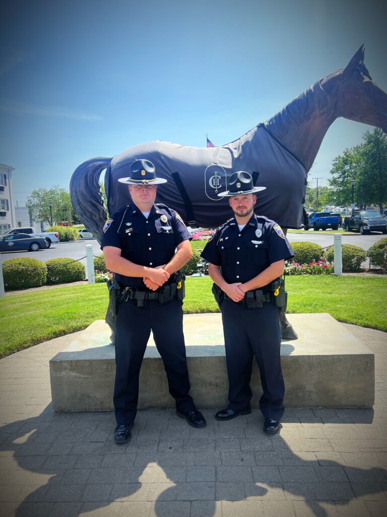“Two uniformed MPD officers in campaign hats stand in front of the covered horse statue on a sunny lawn, one taller than the other, both facing the camera.”