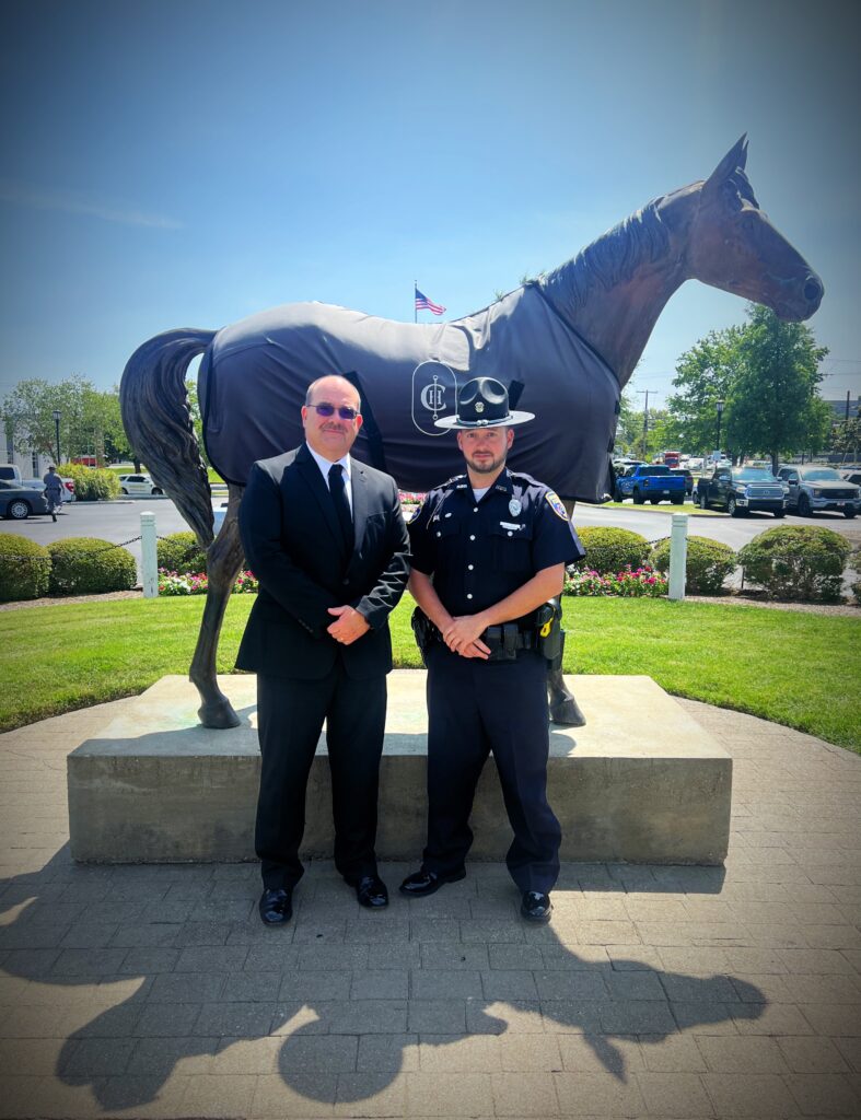 “A suited man and a uniformed MPD officer in campaign hat stand side by side in front of the covered horse statue on a sunny lawn.”