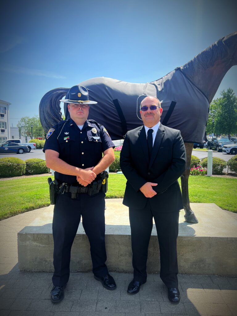 “Uniformed MPD officer in campaign hat and a suited man stand side by side in front of the covered horse statue on a sunny lawn, both facing the camera.”