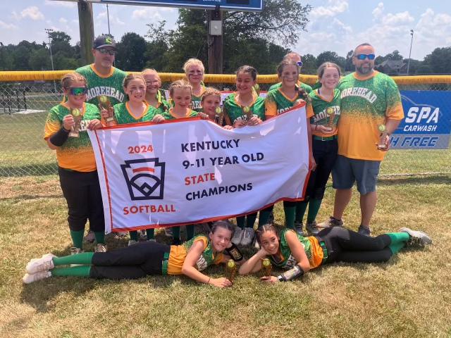 “Youth softball team in green-and-yellow uniforms and two coaches hold a banner reading ‘2024 Kentucky 9-11 Year Old State Champions’ on a sunny field.”