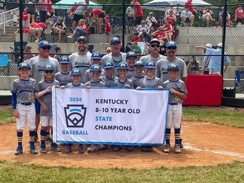 “Youth baseball team in gray-and-blue uniforms and their coaches hold a banner reading ‘2024 Kentucky 8-10 Year Old State Champions’ on a sunny diamond.”